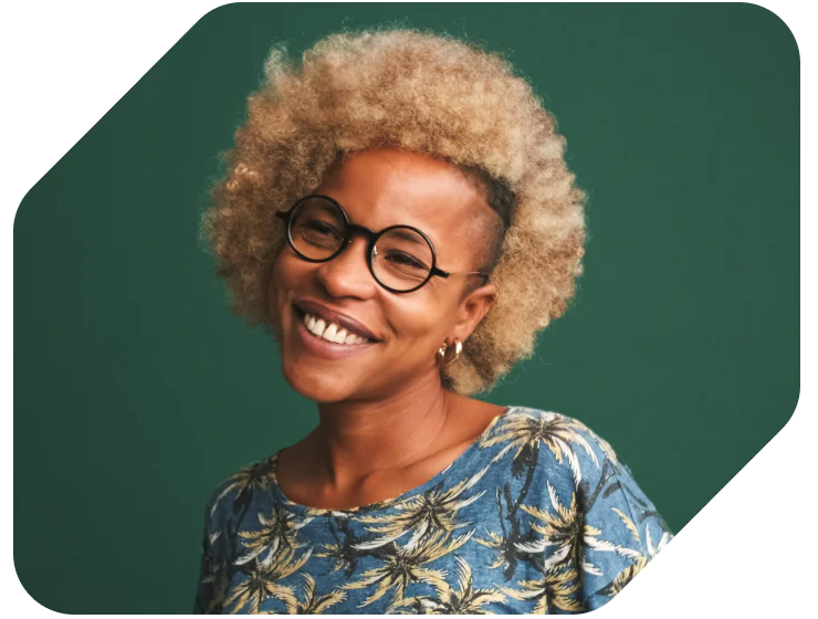 Smiling Person with Afro and Round Glasses Against Green Background Person with blond Afro hairstyle and round glasses, smiling and wearing a blue patterned shirt.